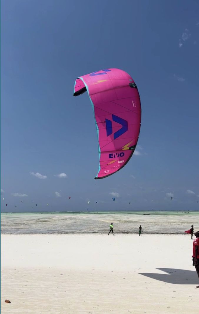 A pink kite flying over the white beach and the lagoon at Paje, with kitesurfers in the distance